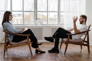Two people in conversation, sitting on chairs indoors.